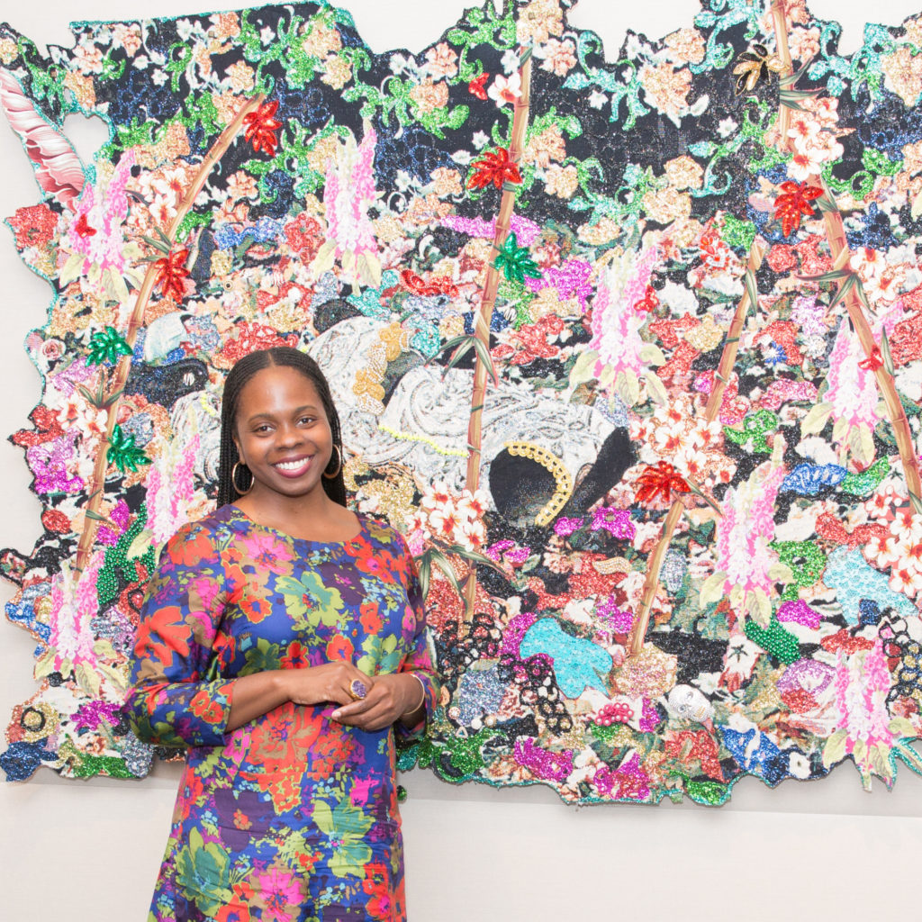 A woman smiles for a photo while posing in front of a large, colorful pink and green work of art on a wall