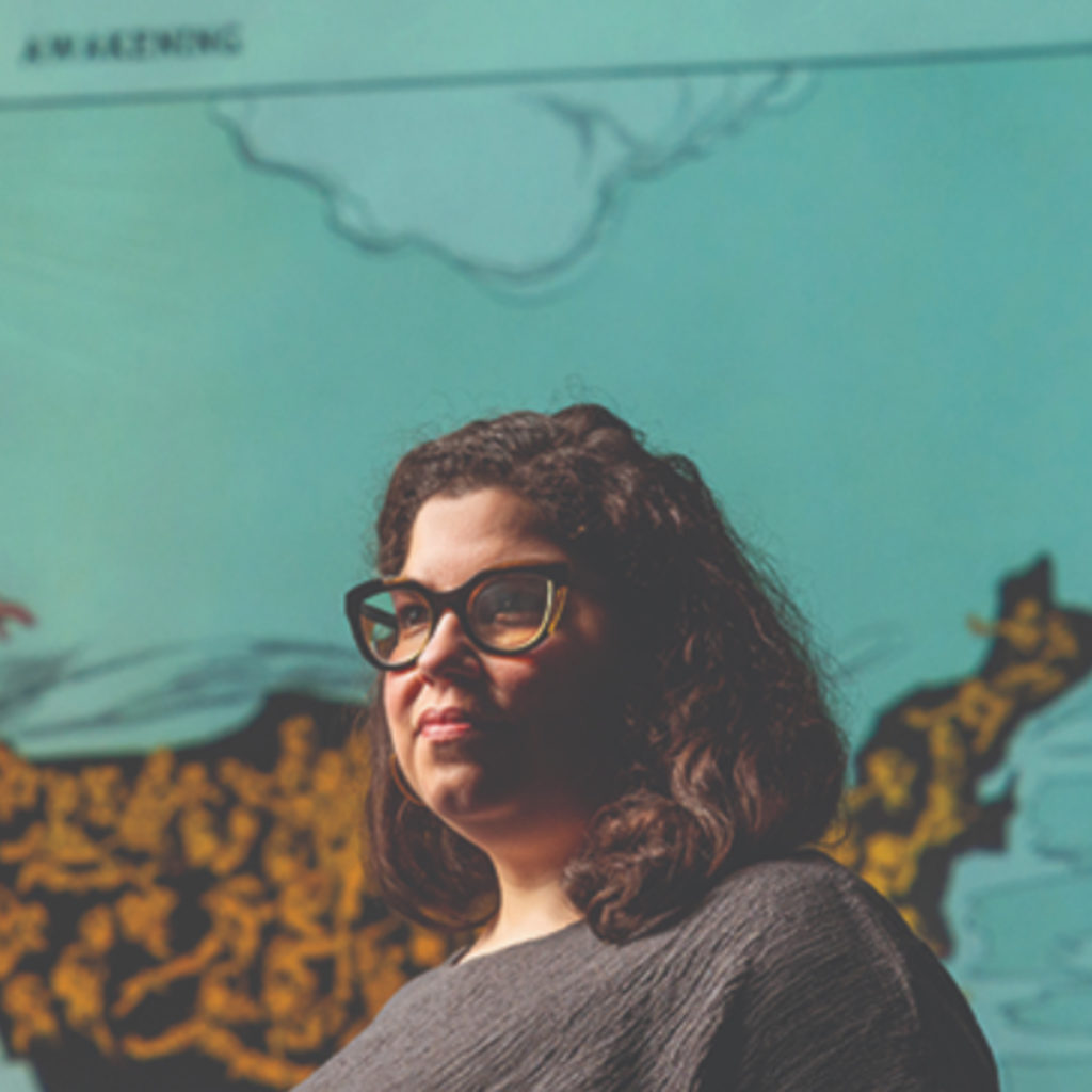 A woman with glasses and shoulder length hair poses in front of an image of a map