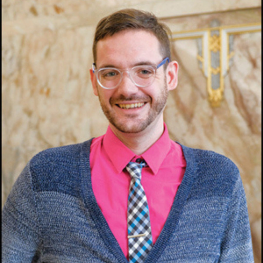 A man in a pink collared shirt and blue cardigan smiles for a portrait