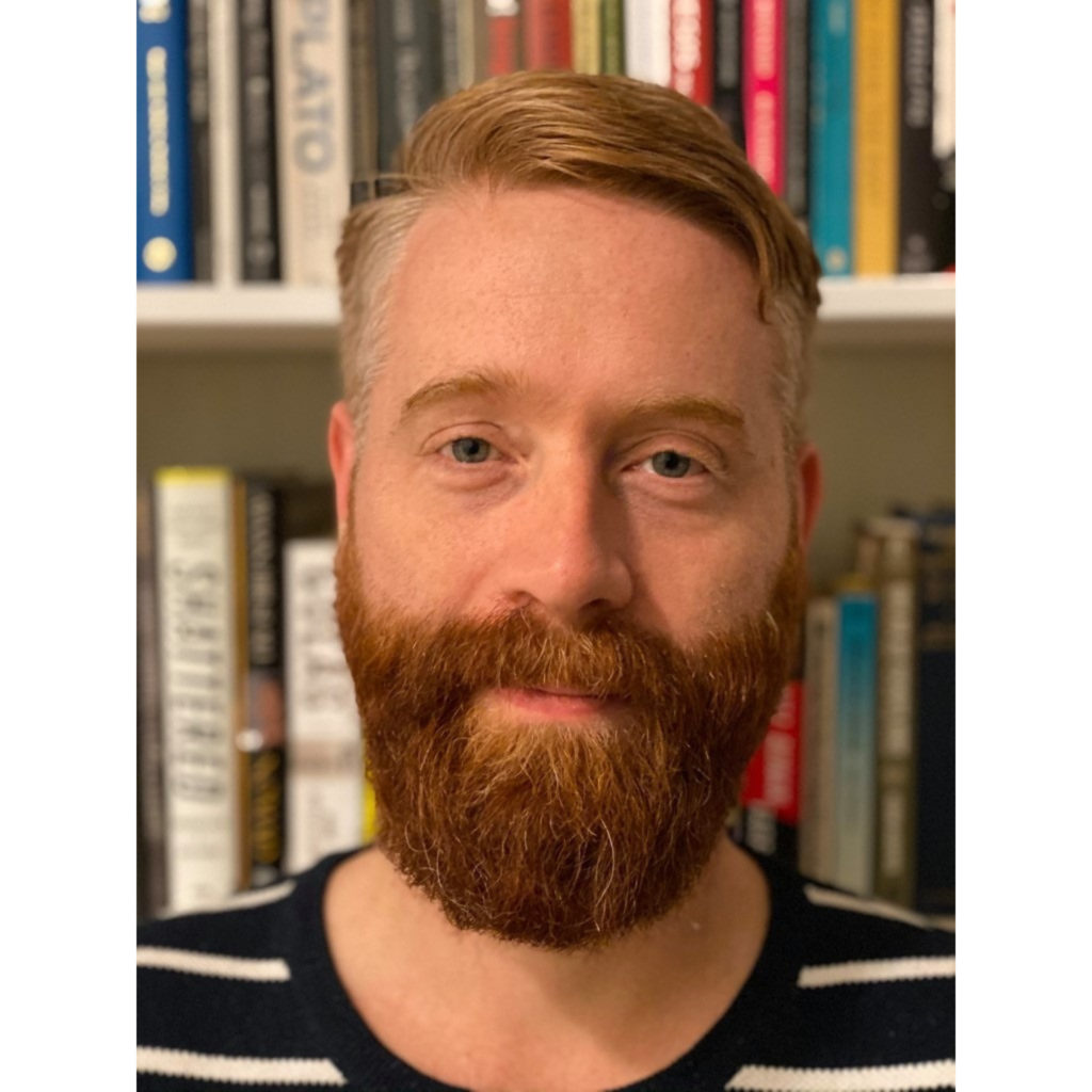 A man with a beard poses for a photo in front of a bookshelf